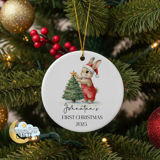 Round ceramic Christmas ornament with bunny in Santa hat placing a star on a decorated tree, personalized with name and year for baby’s first Christmas.