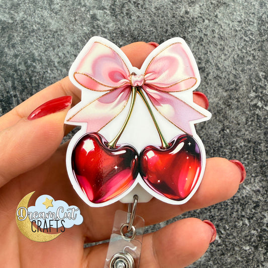 Cherry bow badge reel top, UV printed on 3 mm white acrylic, red cherries with pink bow design.