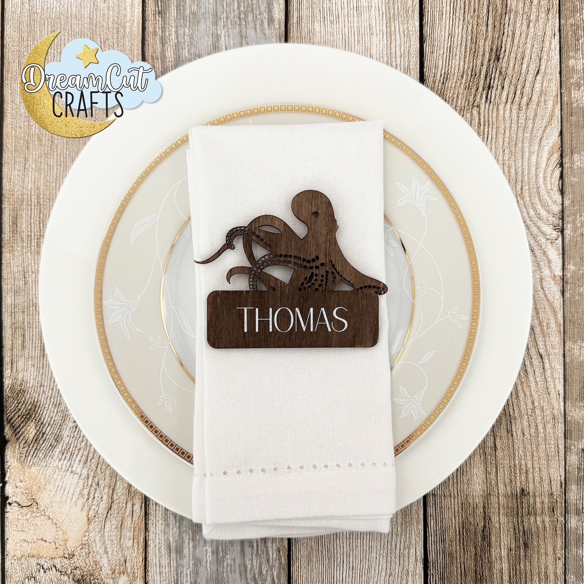 Wooden place card with custom name on a white napkin over a plate, on a wooden surface with 'DreamCrafts' logo.