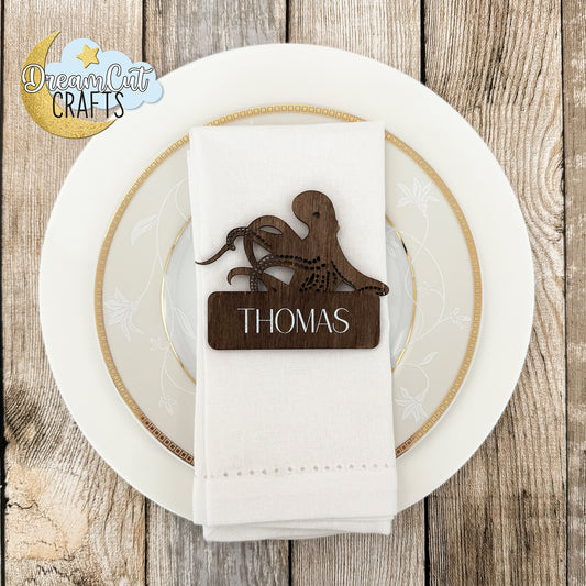 Wooden place card with custom name on a white napkin over a plate, on a wooden surface with 'DreamCrafts' logo.
