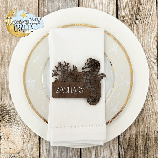Wooden place card with seahorse design on a white plate with gold rim on a wooden surface, featuring 'DreamCrafts' logo.